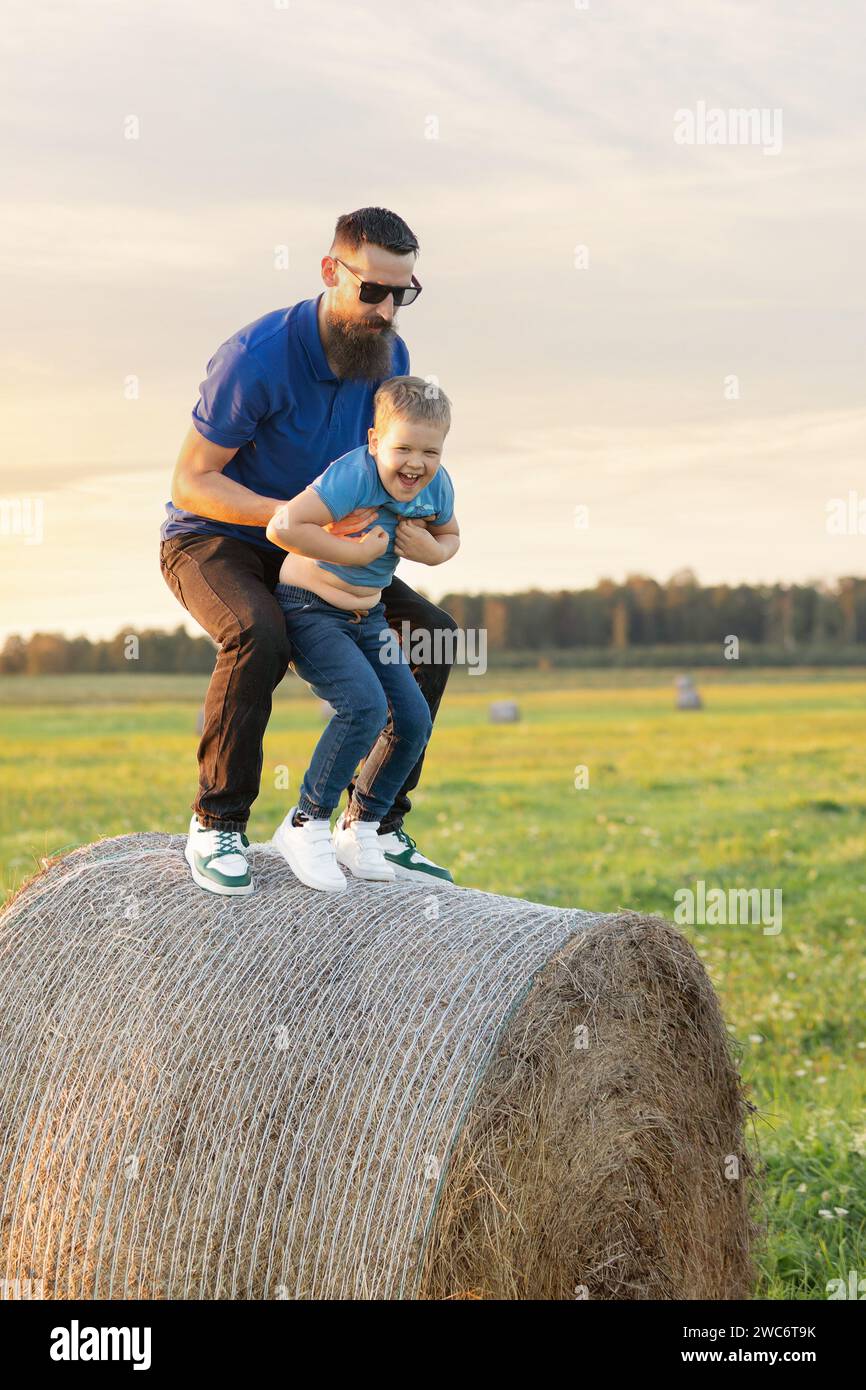 Portrait of a father with his son in an evening sunlight nature background. They play pranks on a hay roll. Stock Photo