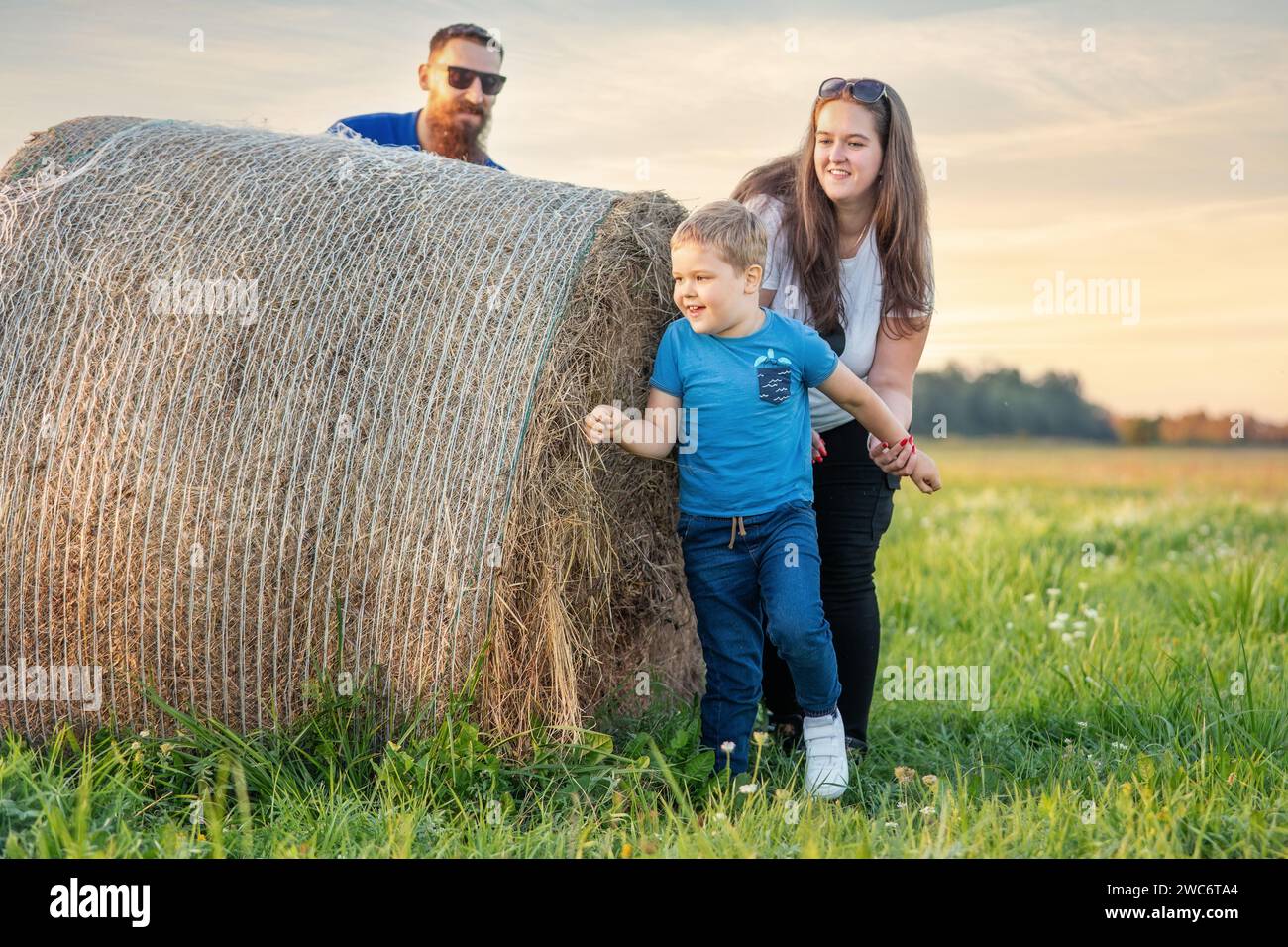 Summer holiday evening fun family games of hide and seek in the meadow around the hay bale. Mother and son catching father and he hides behind a roll Stock Photo