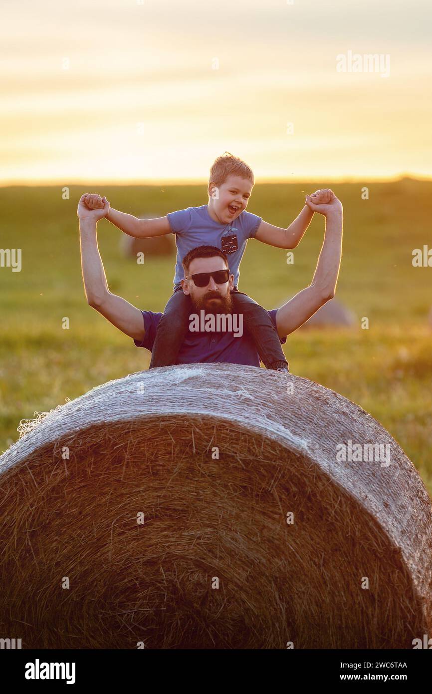 Portrait of a father holding his son on his shoulders. The strong emotion of a happy child. Holding hands. A roll of hay in the foreground. Stock Photo