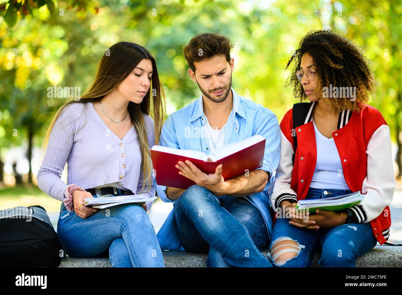 Three multiethnic students studying together sitting on a bench outdoor Stock Photo