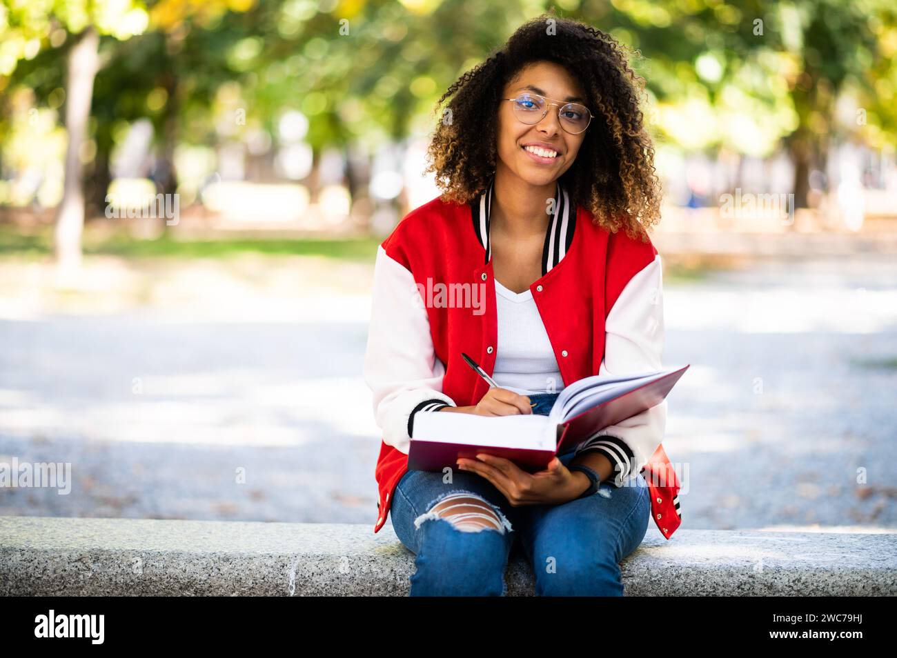 Beautiful african female college student reading a book on a bench in a park Stock Photo