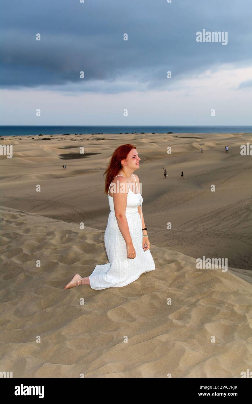 A young girl kneels sadly in a white dress in the sand dunes under an overcast sky Stock Photo