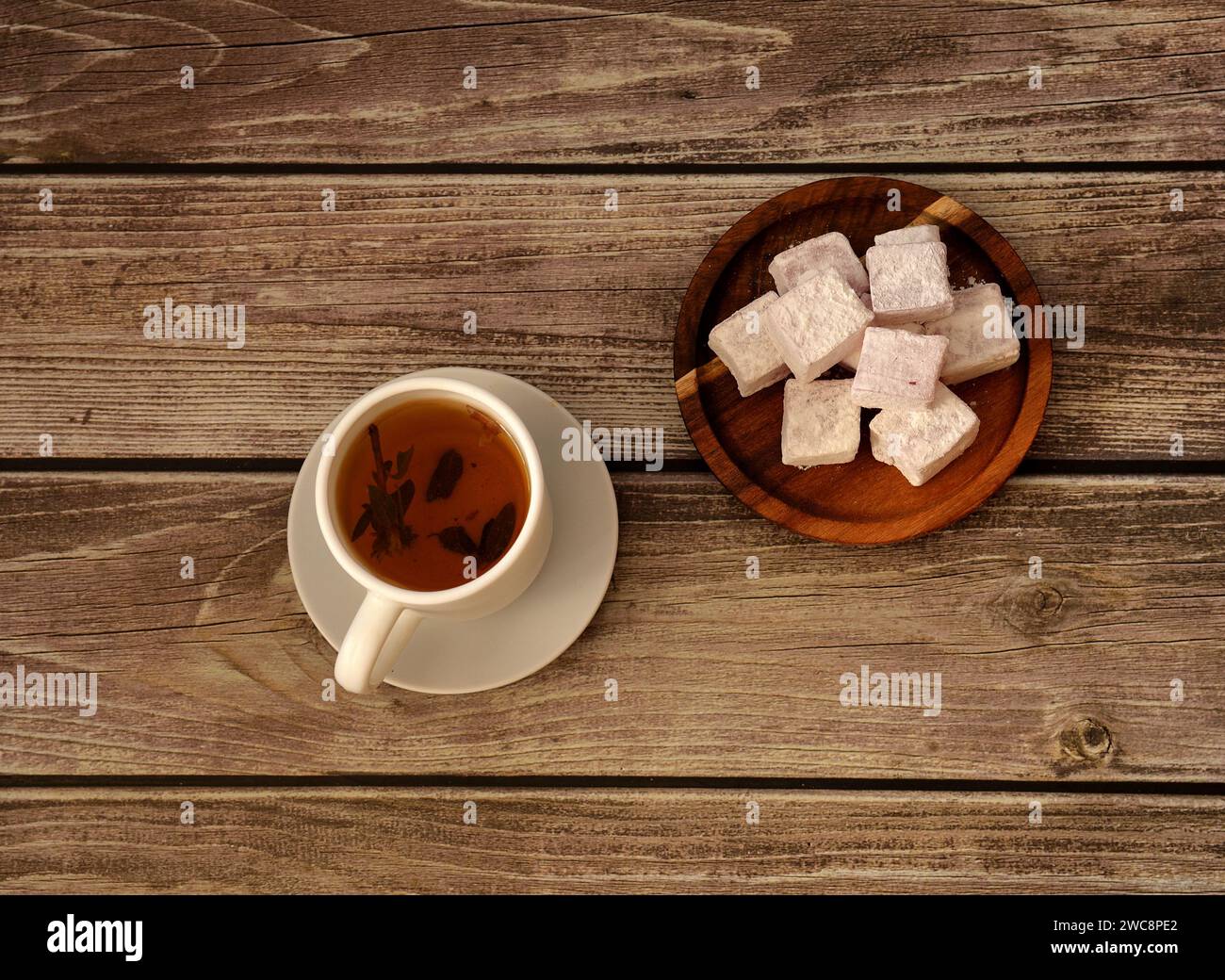 A cup of hot herbal tea on a saucer and a round wooden plate with a bunch of oriental sweets in powdered sugar on a wooden table. Top view, flat lay. Stock Photo