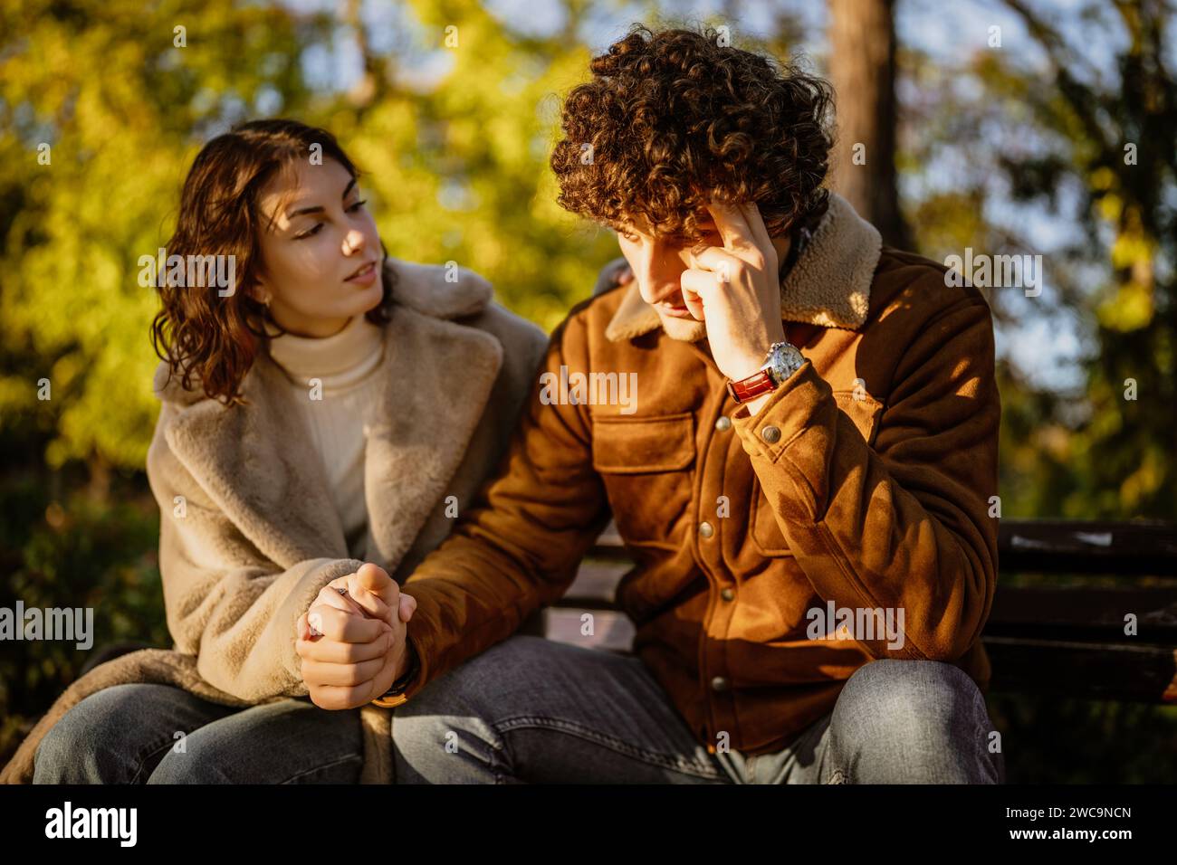 Young couple is sitting in park on sunny day. Man is sad and woman in consoling him. Stock Photo