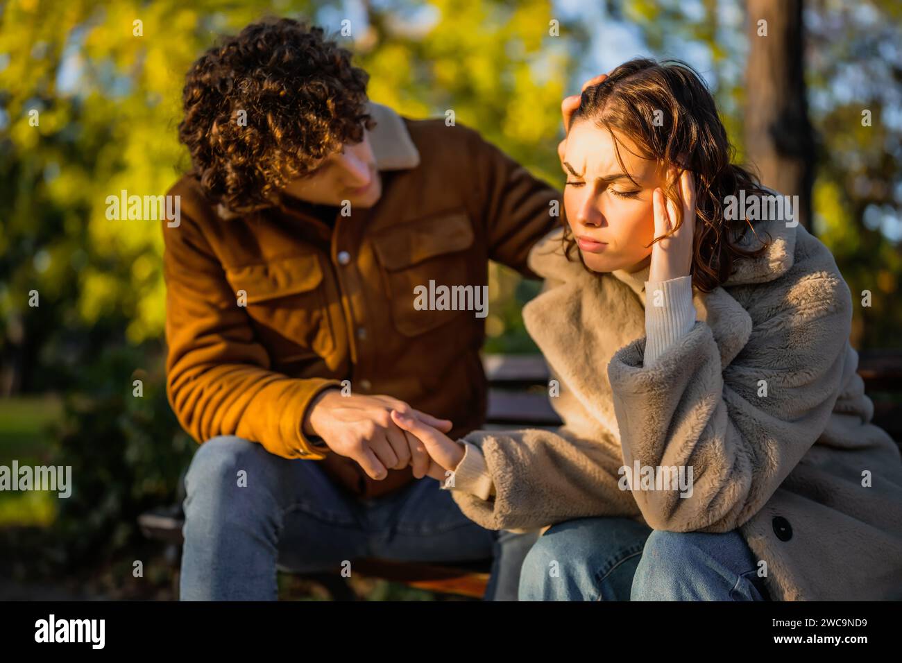 Young couple is sitting in park on sunny day. Woman is sad and man in consoling her. Stock Photo