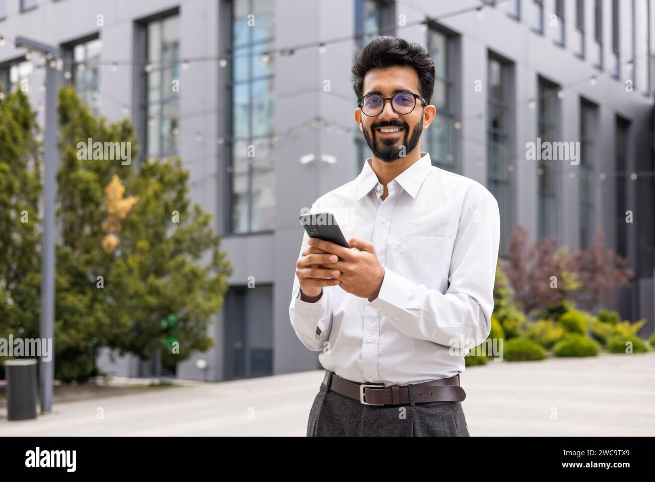 Confident young businessman with a beard, smiling while using smartphone outdoors, office building backdrop. Stock Photo