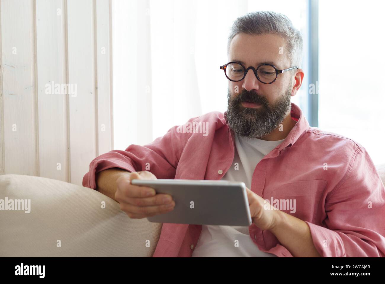 Man using smart thermostat, adjusting, lowering heating temperature at home. Man watching tv show on tablet, video streaming services with movies and Stock Photo