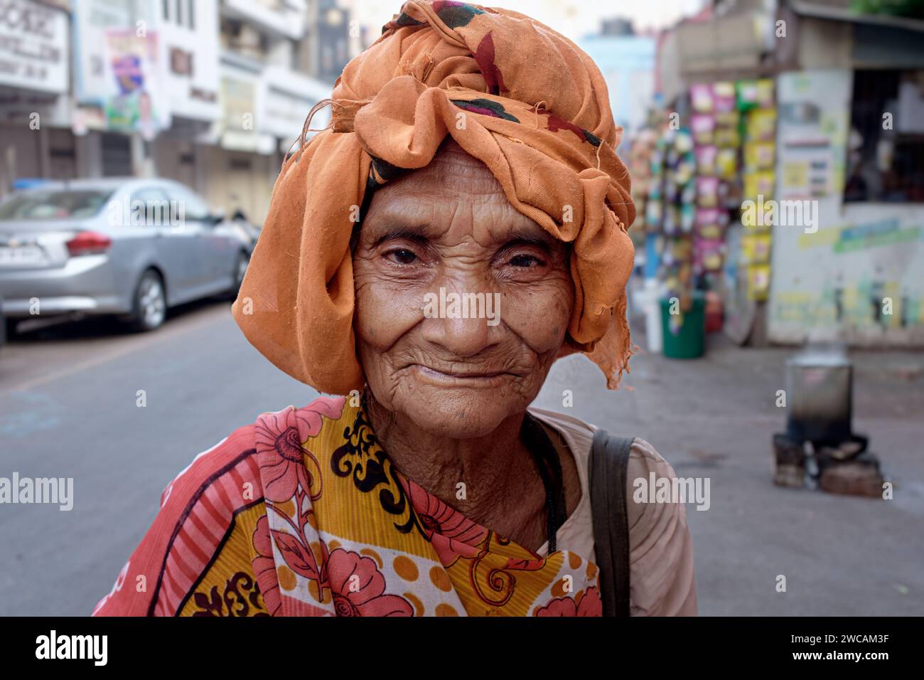 An old, homeless woman outside Mumbadevi Temple, Mumbai, India, her orange or saffron-colored head dress indicating Hindu faith, surviving by begging Stock Photo