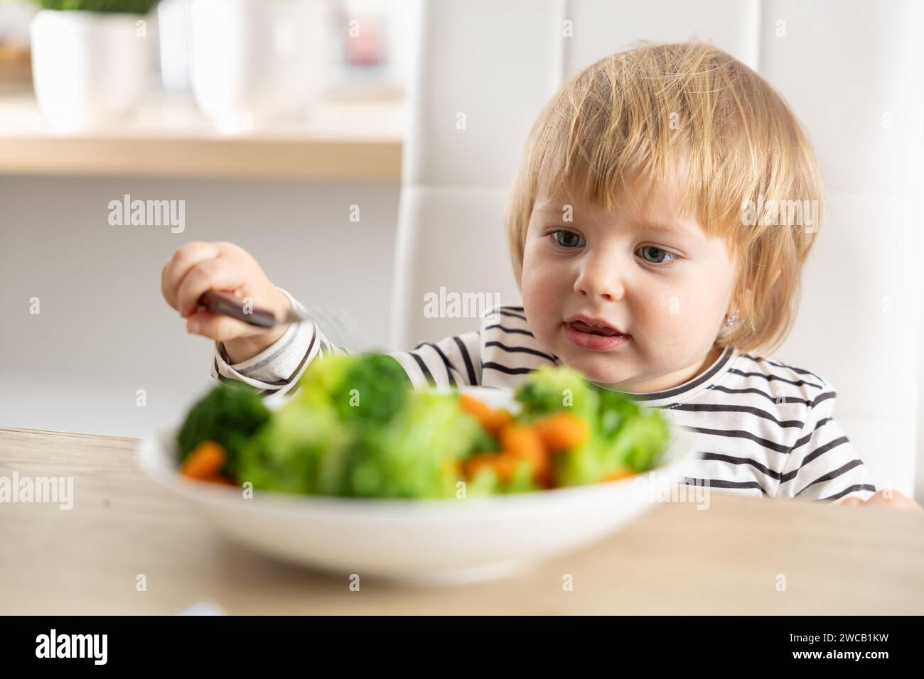 Cute little girl is eating broccoli and carrot vegetables with a fork