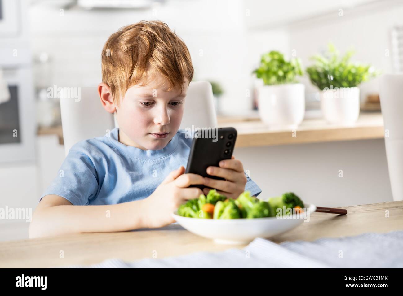 View of smart cute boy using smartphone before lunch in kitchen at home. Stock Photo