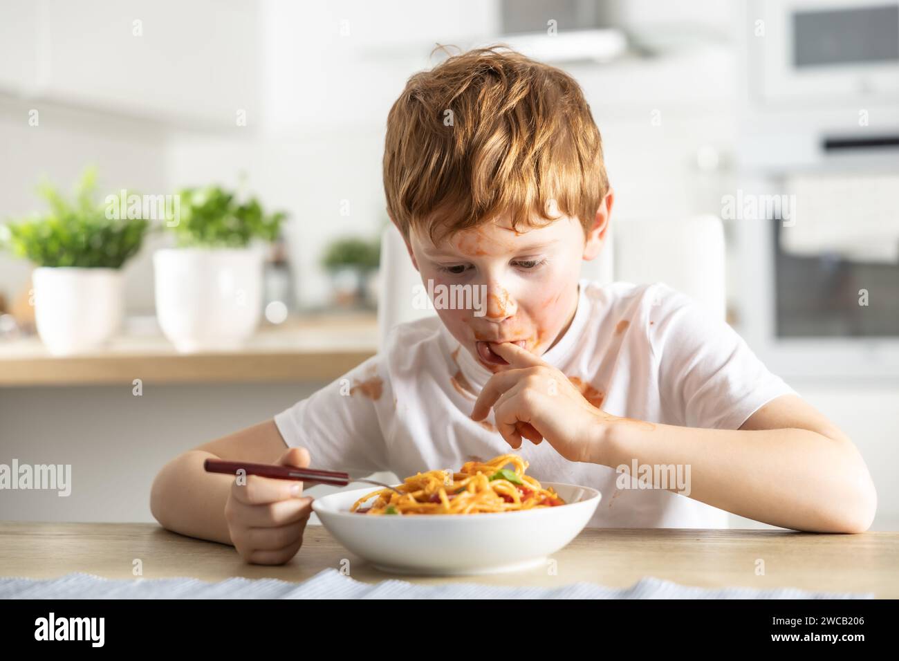 A cute little boy is eating spaghetti bolognese for lunch in the kitchen at home and is covered in ketchup and licking his fingers. Stock Photo