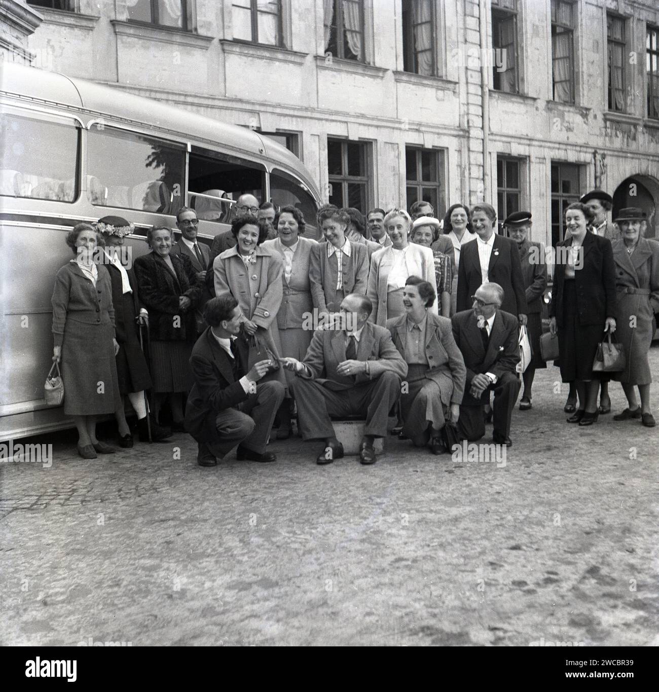 1950, historical, in a cobbled Frrench courtyard, a group of adults gathered outside for photo by their coach of their era, a Dennis. The men and women are on a trip visiting Commonwealth War graves and wearing formal clothes.. lots of laughter. Stock Photo