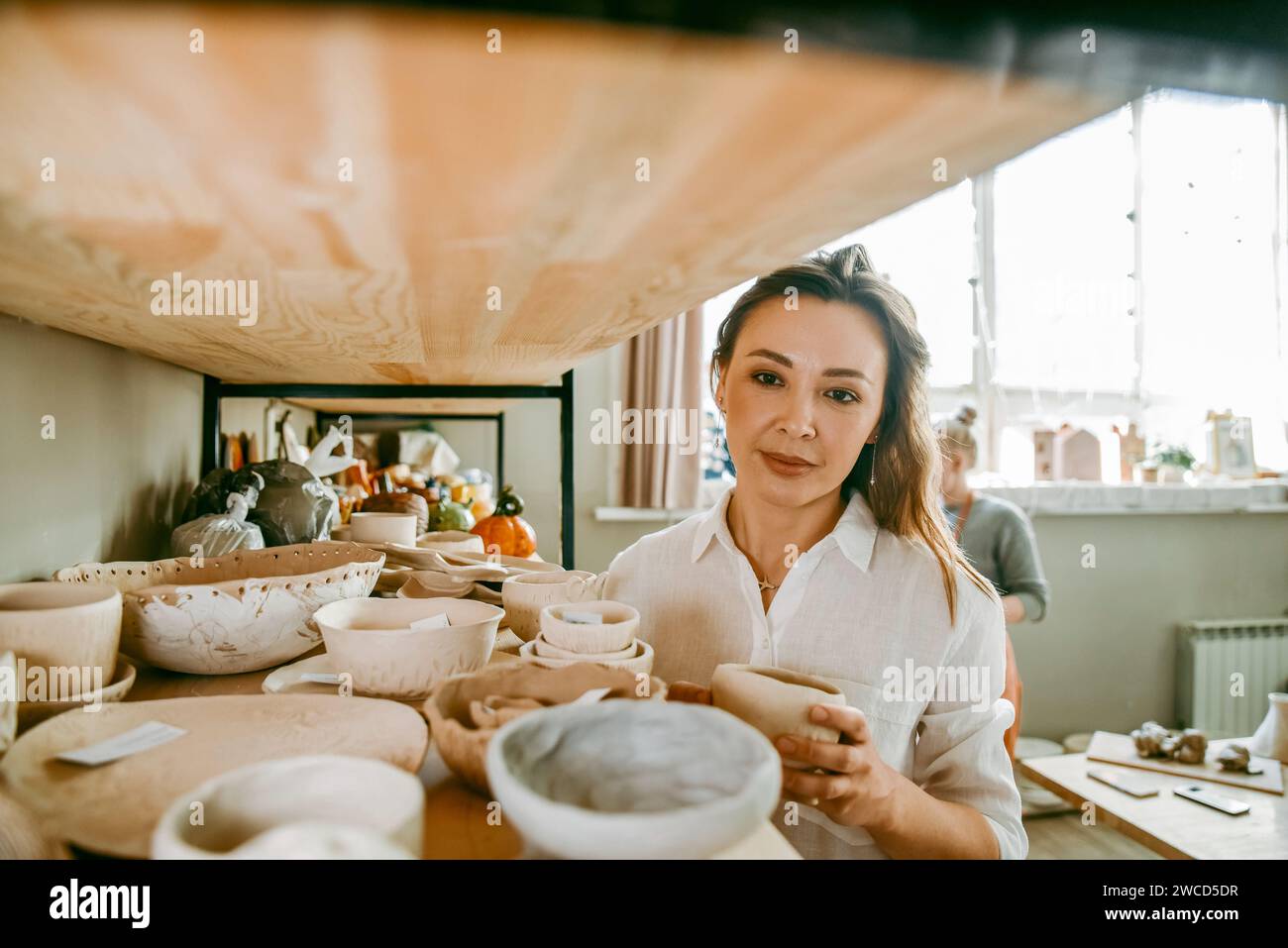 A beautiful middle-aged woman stands at shelf with various pottery items. A young woman presents clay products in her shop-workshop, small business. Stock Photo