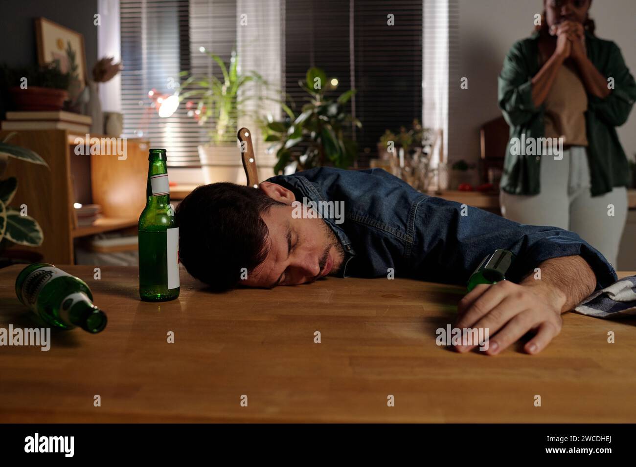 Young drunk man with knife in his neck keeping head on wooden kitchen table with group of empty beer bottles against his wife Stock Photo