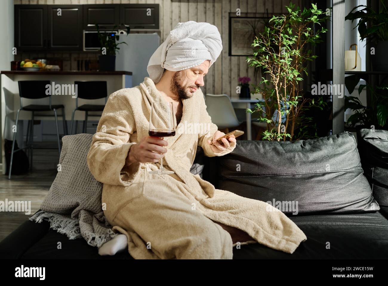 Young relaxed man in bathrobe holding glass of red wine and texting in smartphone or watching online video while sitting on couch Stock Photo
