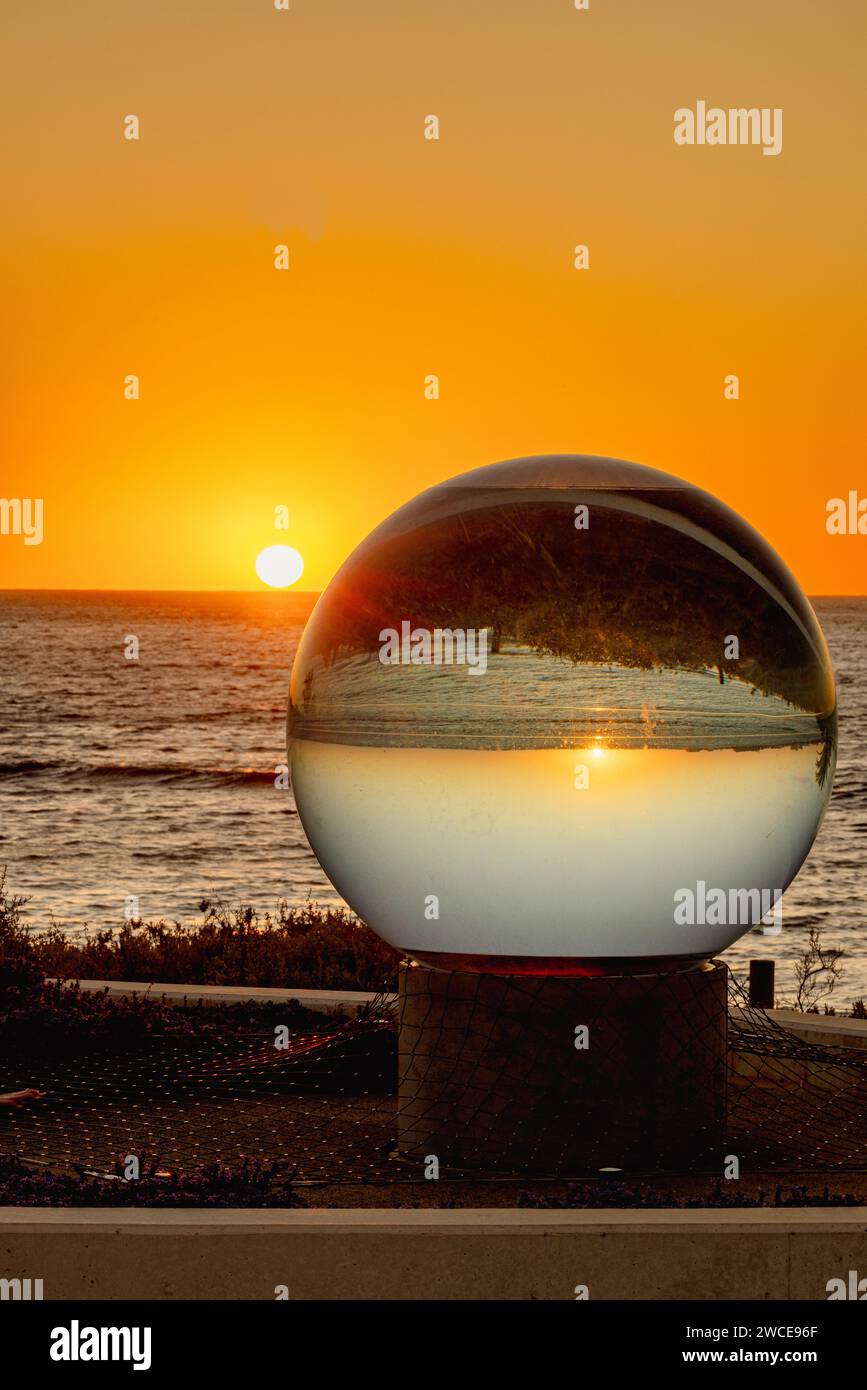 A transparent glass sphere rests on the water's edge as the sun sets in the background, creating a captivating and serene scene Stock Photo
