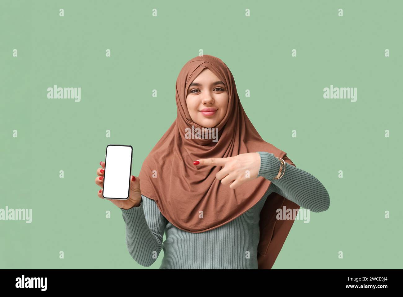 Beautiful young Muslim woman in hijab pointing at mobile phone on green ...