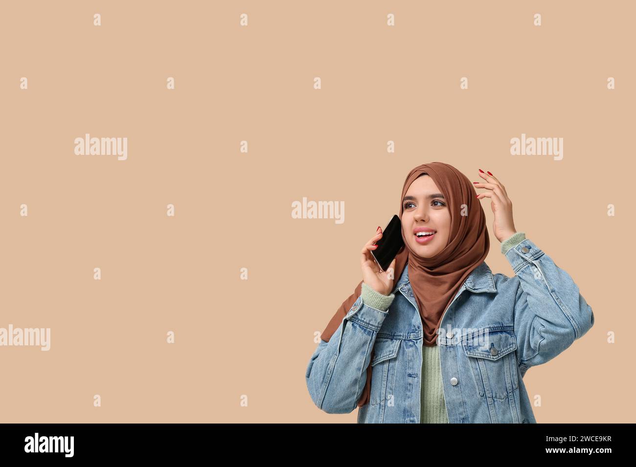Beautiful young Muslim woman in hijab talking by mobile phone on beige ...