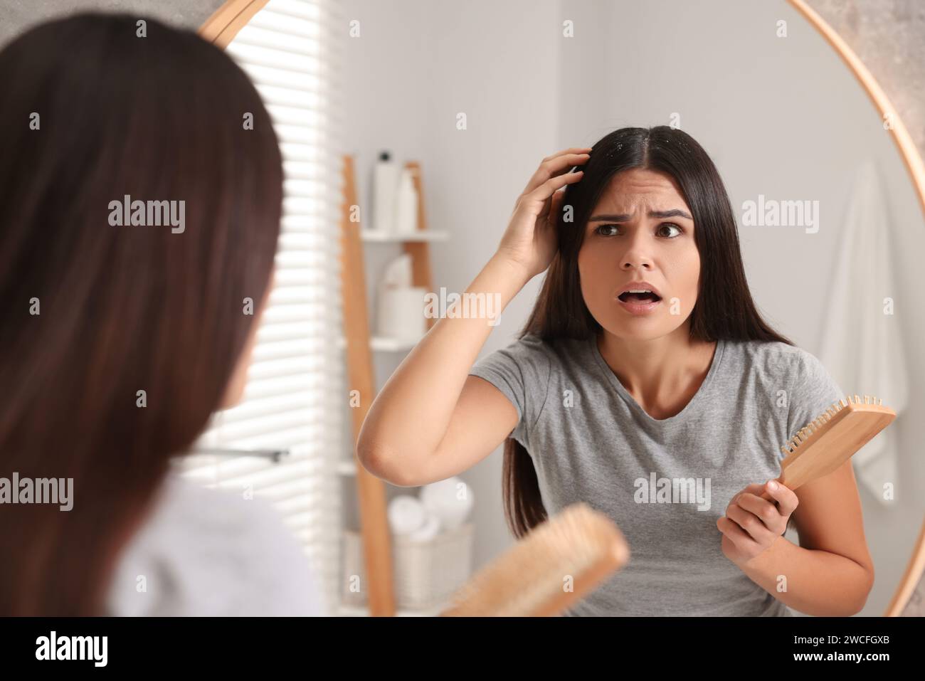 Emotional woman with brush examining her hair and scalp near mirror in bathroom. Dandruff problem Stock Photo