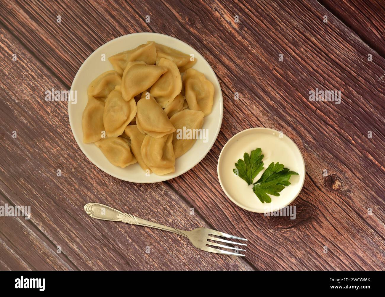 A plate of freshly cooked dumplings on a wooden table, next to a fork and a cup of sour cream with herbs. Close-up. Stock Photo