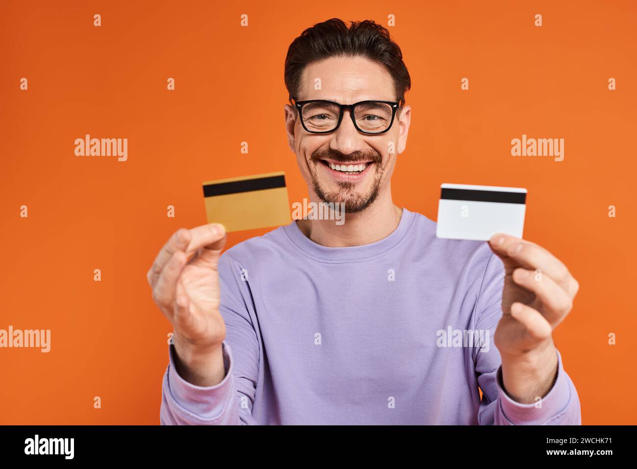 cheerful bearded man in glasses holding credit cards on orange background, shopping and consumerism Stock Photo