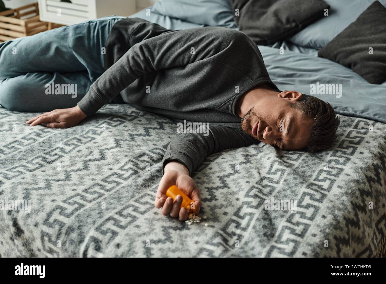 bearded man suffering from pain and looking at bottle with pills while lying on bed with blanket Stock Photo