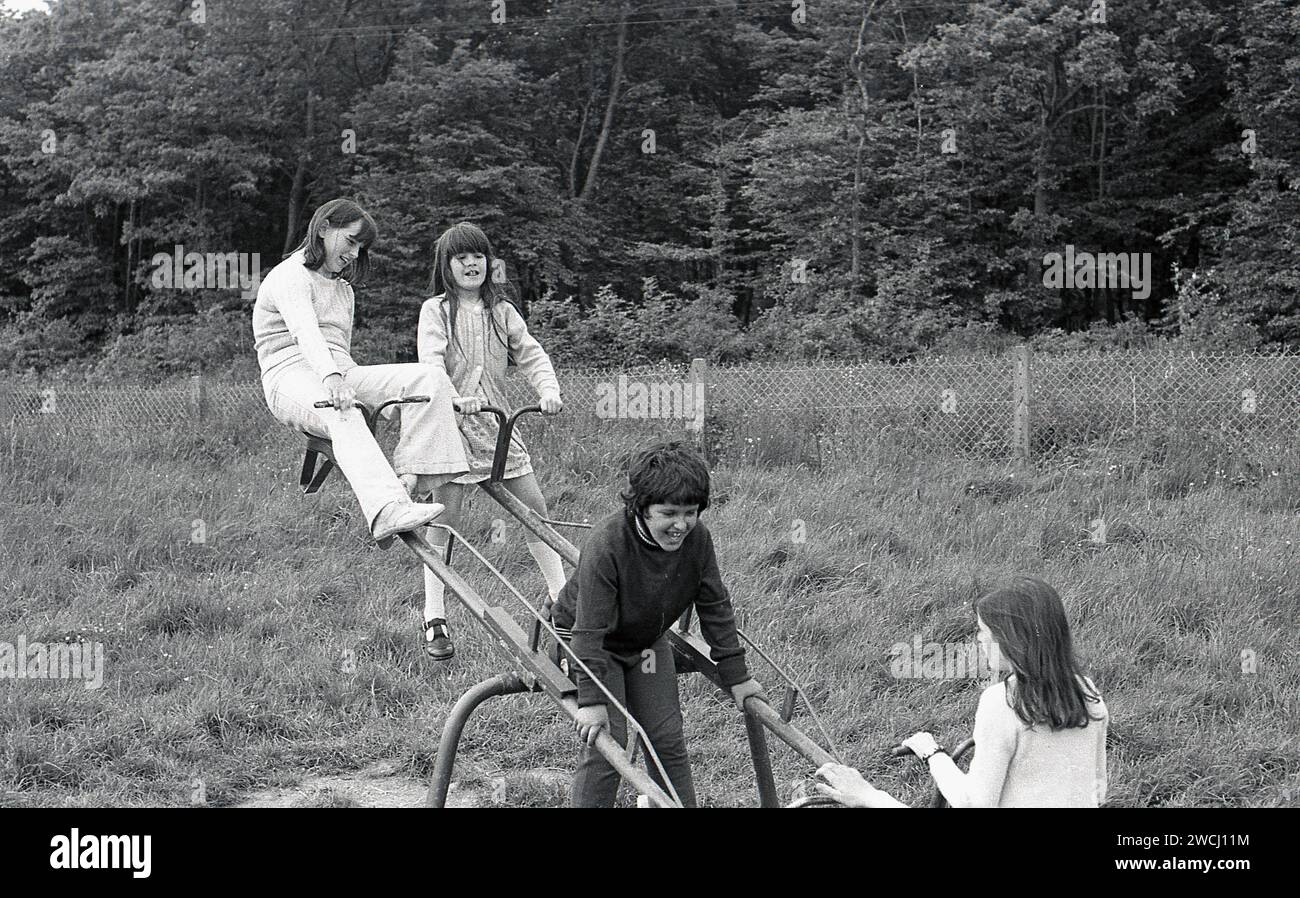 1970s, historical, in a grassy field beside woodland, youngsters playing outside having fun together riding on a double metal see-saw, Essex, England, UK. Despite them being great fun and a healthy activity for growing boys and girls, See-saws became a less common sight in British playgrounds due to safety concerns. Stock Photo