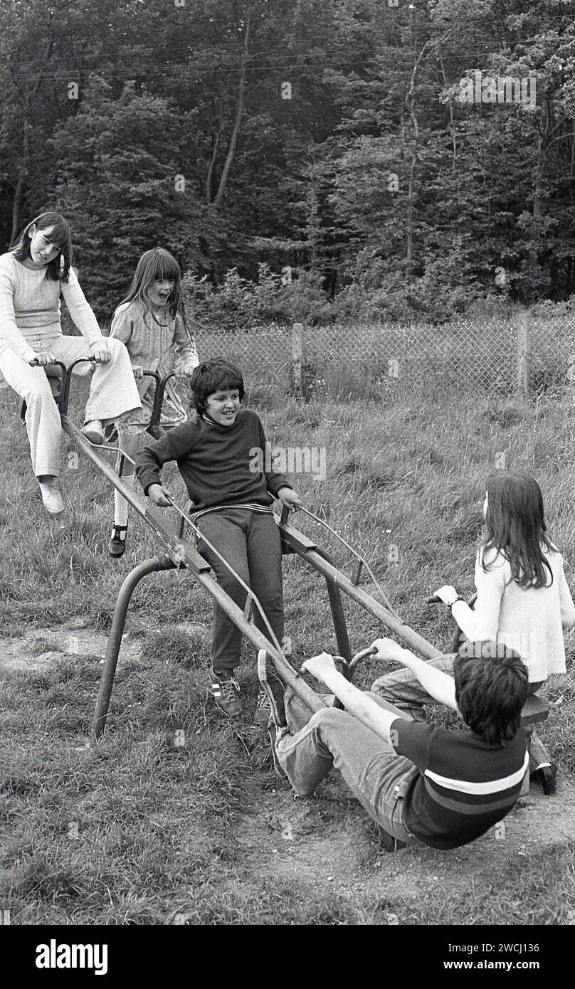 1970s, historical, in a grassy field beside woodland, youngsters playing outside having fun together riding on a double metal see-saw, Essex, England, UK. Despite them being great fun and a healthy activity for growing boys and girls, See-saws became a less common sight in British playgrounds due to safety concerns. Stock Photo