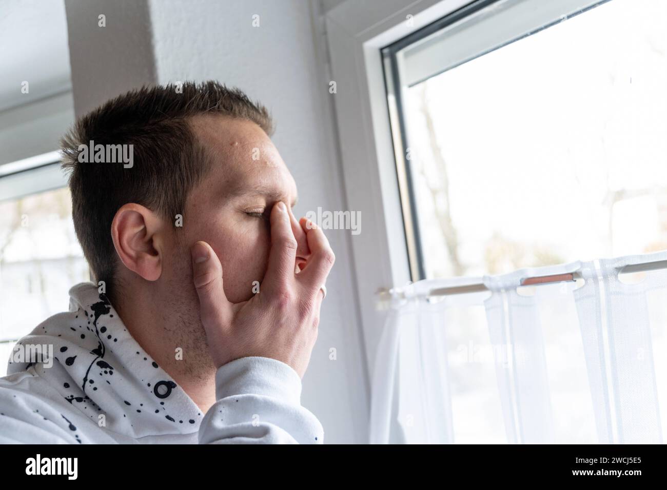 Augsburg, Bavaria, Germany - January 14, 2024: Young man stands sad and desperate at a window, holding his face in pain *** Junger Mann steht traurig und verzweifelt an einem Fenster, er hält sich das Gesicht vor schmerz Stock Photo