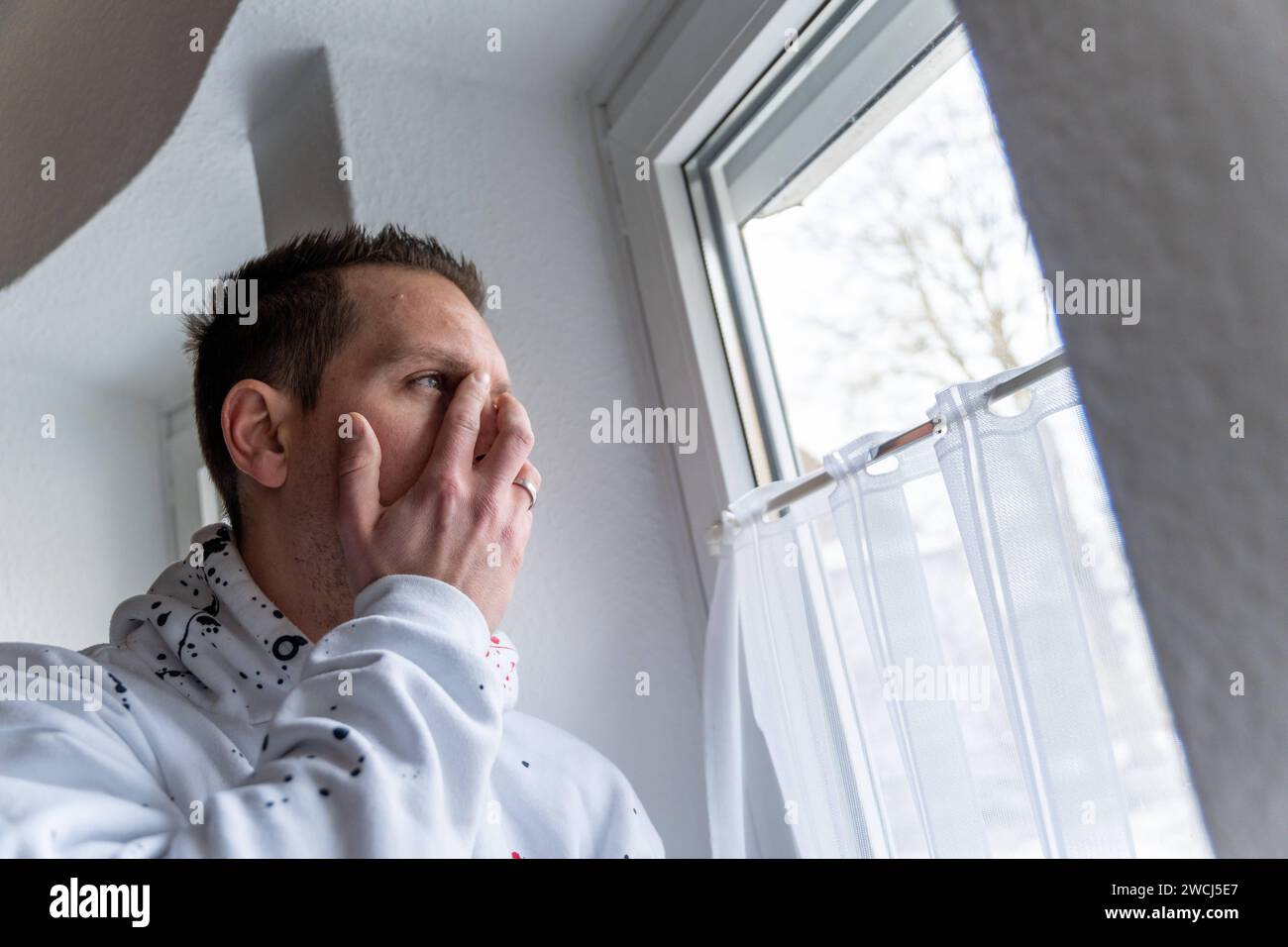 Augsburg, Bavaria, Germany - January 14, 2024: Young man stands sad and desperate at a window, holding his face in pain *** Junger Mann steht traurig und verzweifelt an einem Fenster, er hält sich das Gesicht vor schmerz Stock Photo