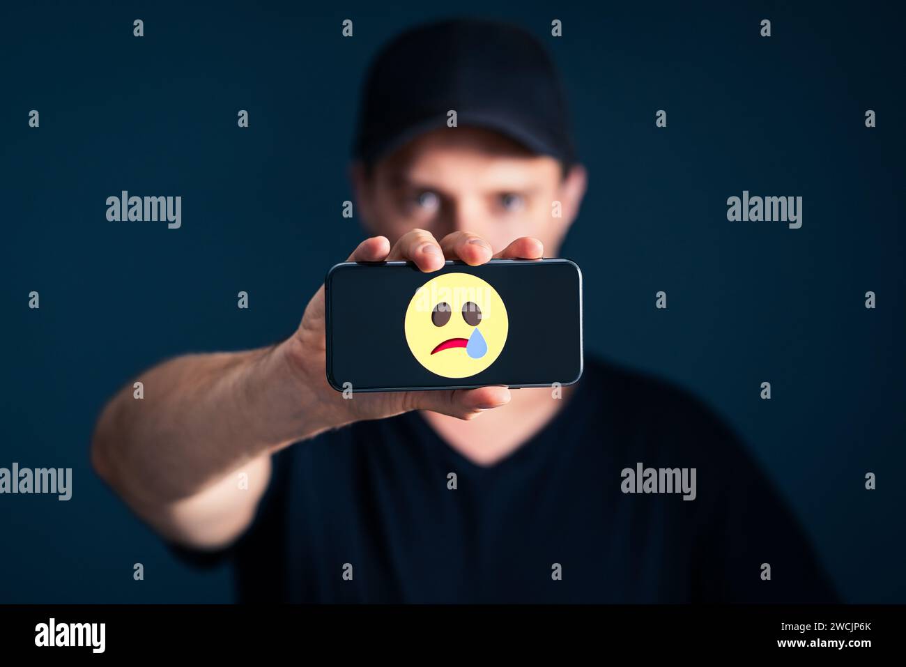 Sad unhappy emotion icon in phone. Depressed lonely man with smartphone. Online hate, trauma, cyber bullying or social media pressure concept. Stock Photo