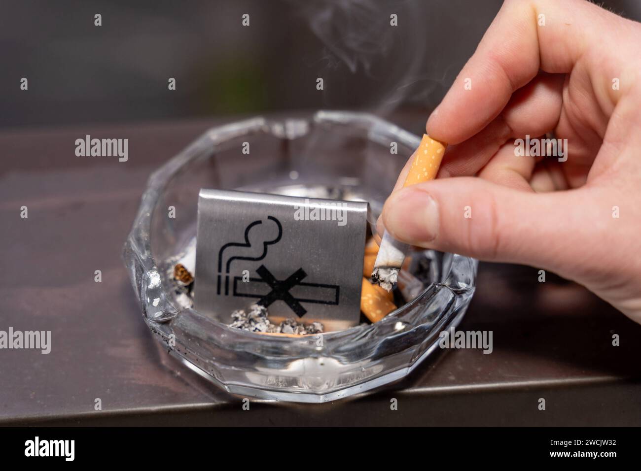 Bavaria, Germany - January 6, 2024: Smoke-free concept, a man puts out a burning cigarette in an ashtray with a no smoking sign *** Rauchfrei Konzept, ein Mann drückt eine brennende Zigarette in einem Aschenbecher mit einem Rauchverbot Schild aus Stock Photo