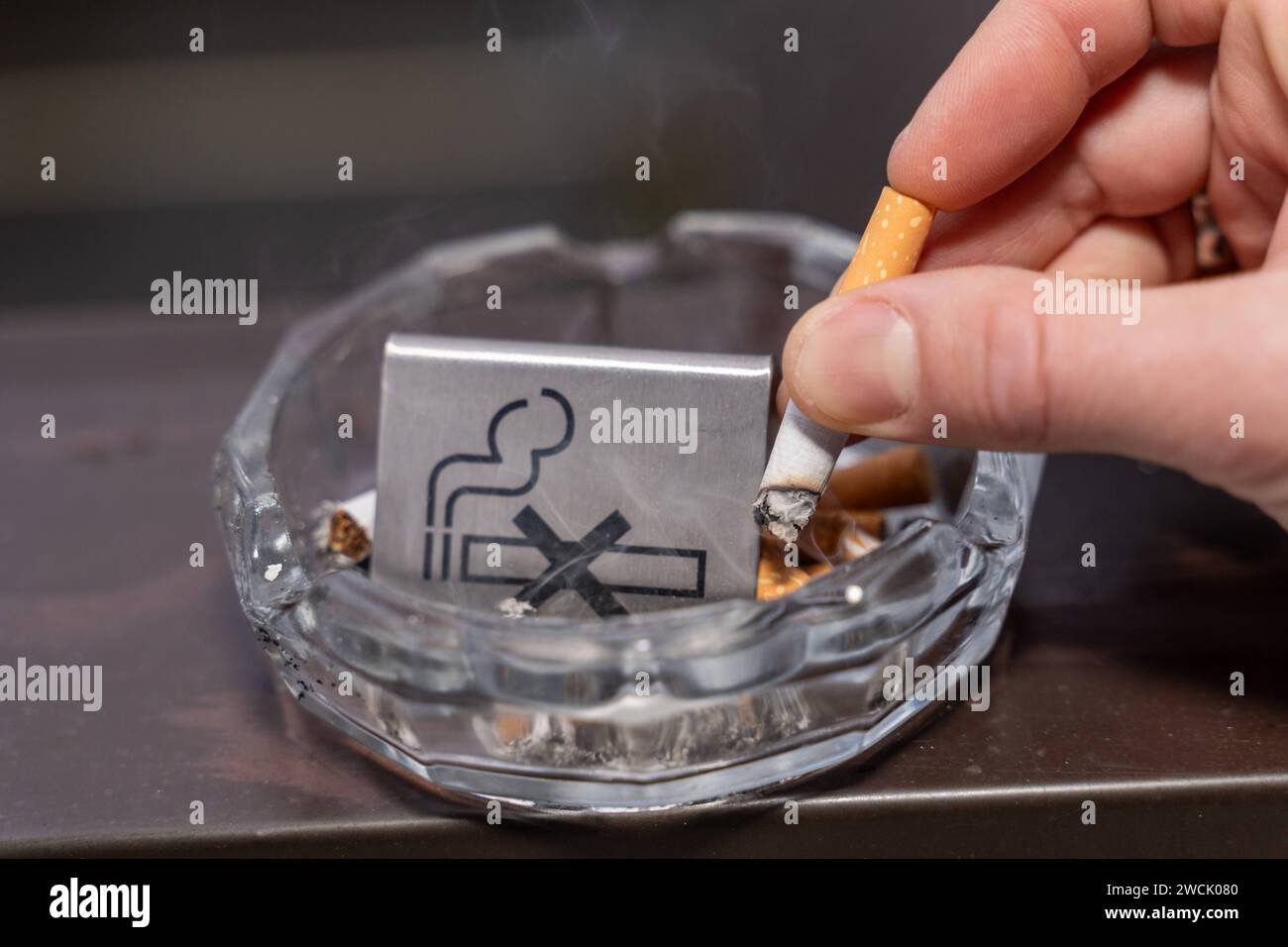 Bavaria, Germany - January 6, 2024: Smoke-free concept, a man puts out a burning cigarette in an ashtray with a no smoking sign *** Rauchfrei Konzept, ein Mann drückt eine brennende Zigarette in einem Aschenbecher mit einem Rauchverbot Schild aus Stock Photo
