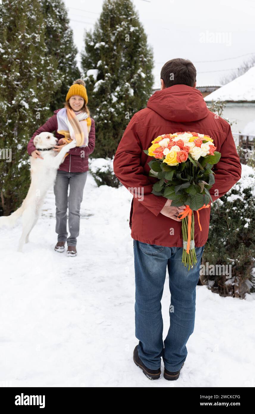 man hides a colorful large bouquet of flowers behind his back, and a happy woman stands in front of him in background, out of focus. Valentine's Day. Stock Photo