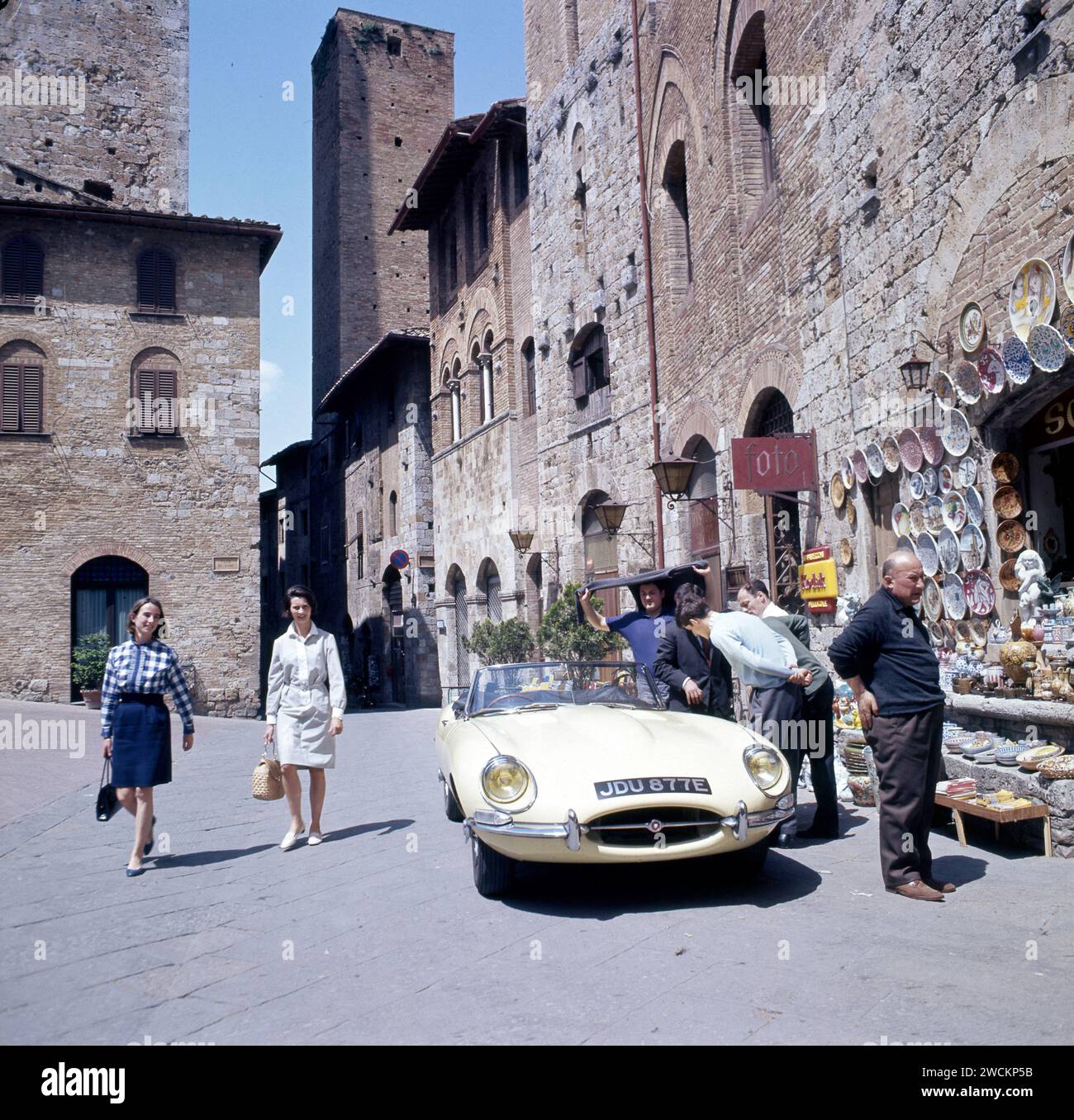 Onlookers admiring Pale Primrose Yellow convertible, Jaguar E-type, JDU877E, parked outside souvenir shop in an Italian square in the summer of 1967. Stock Photo