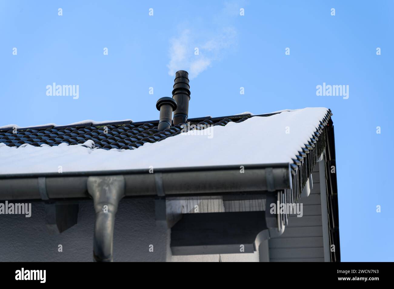 Snow-covered, tiled roof with a chimney from central gas heating unit emitting smoke and water vapor against a clear blue sky. Stock Photo