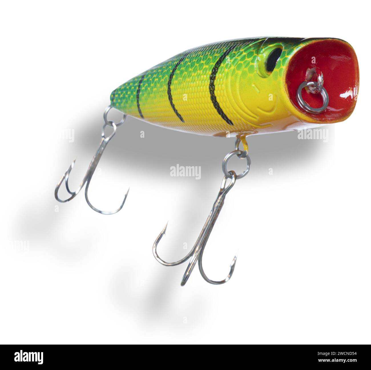 Shadow below a yellow and green artificial topwater fishing bait with red around the eyelet and wearing two treble hooks seen from a steep angle. Stock Photo
