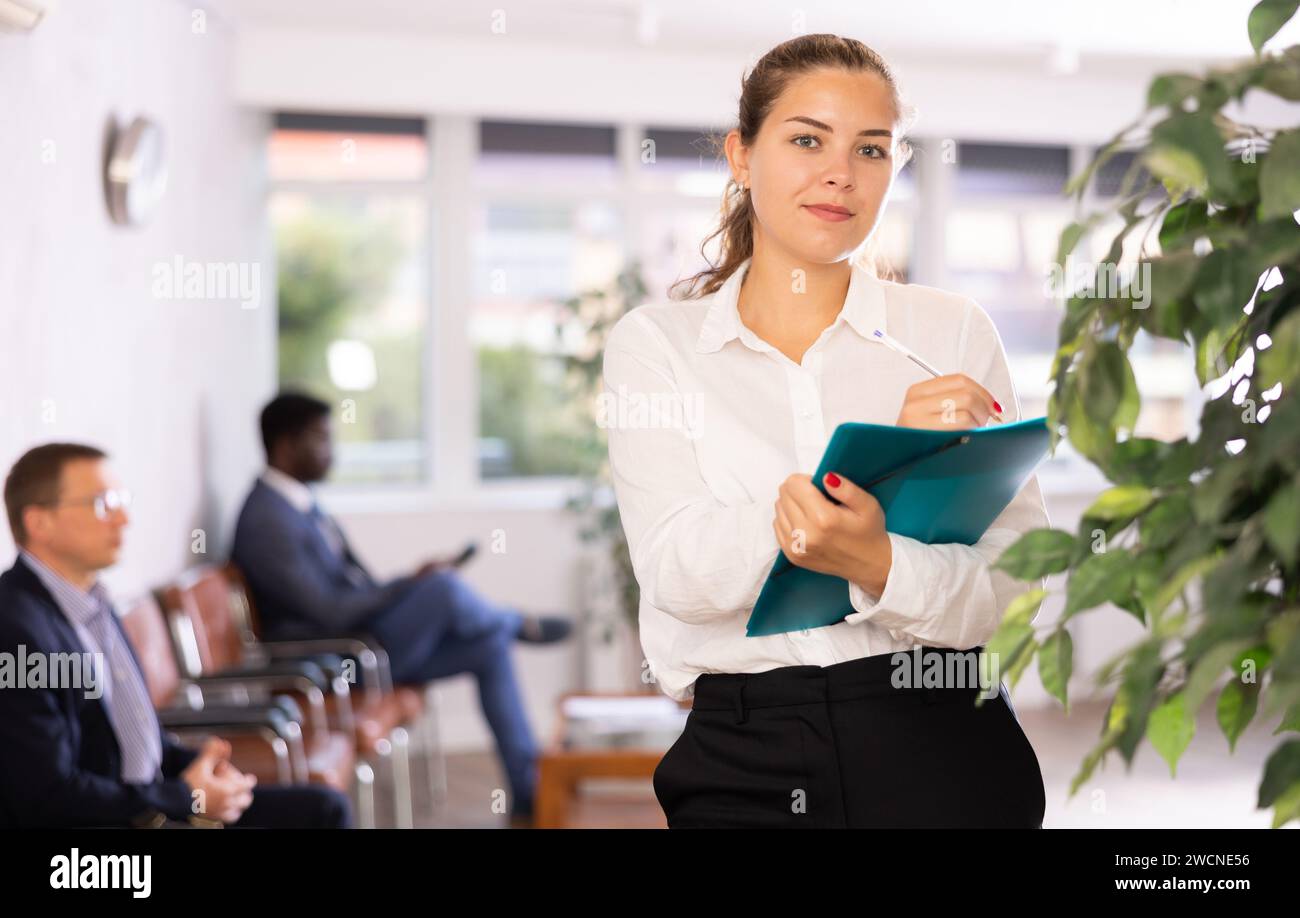 Friendly hostess woman stands at office reception with list of guests and expects invited visitors Stock Photo
