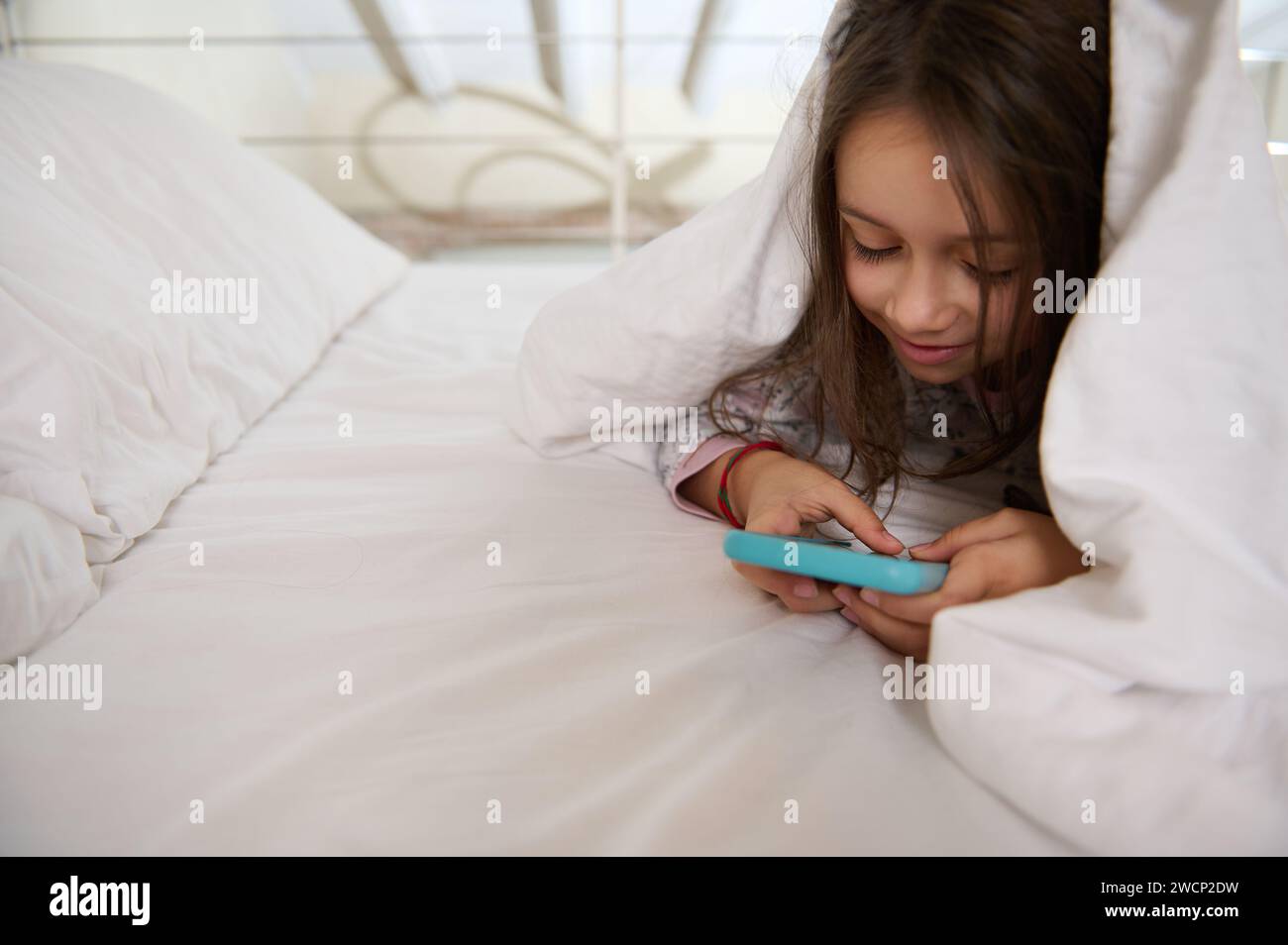 Curious mischievous little kid girl in pajamas, playing online video games, washing cartoons, checking social media content on her smart mobile phone, Stock Photo