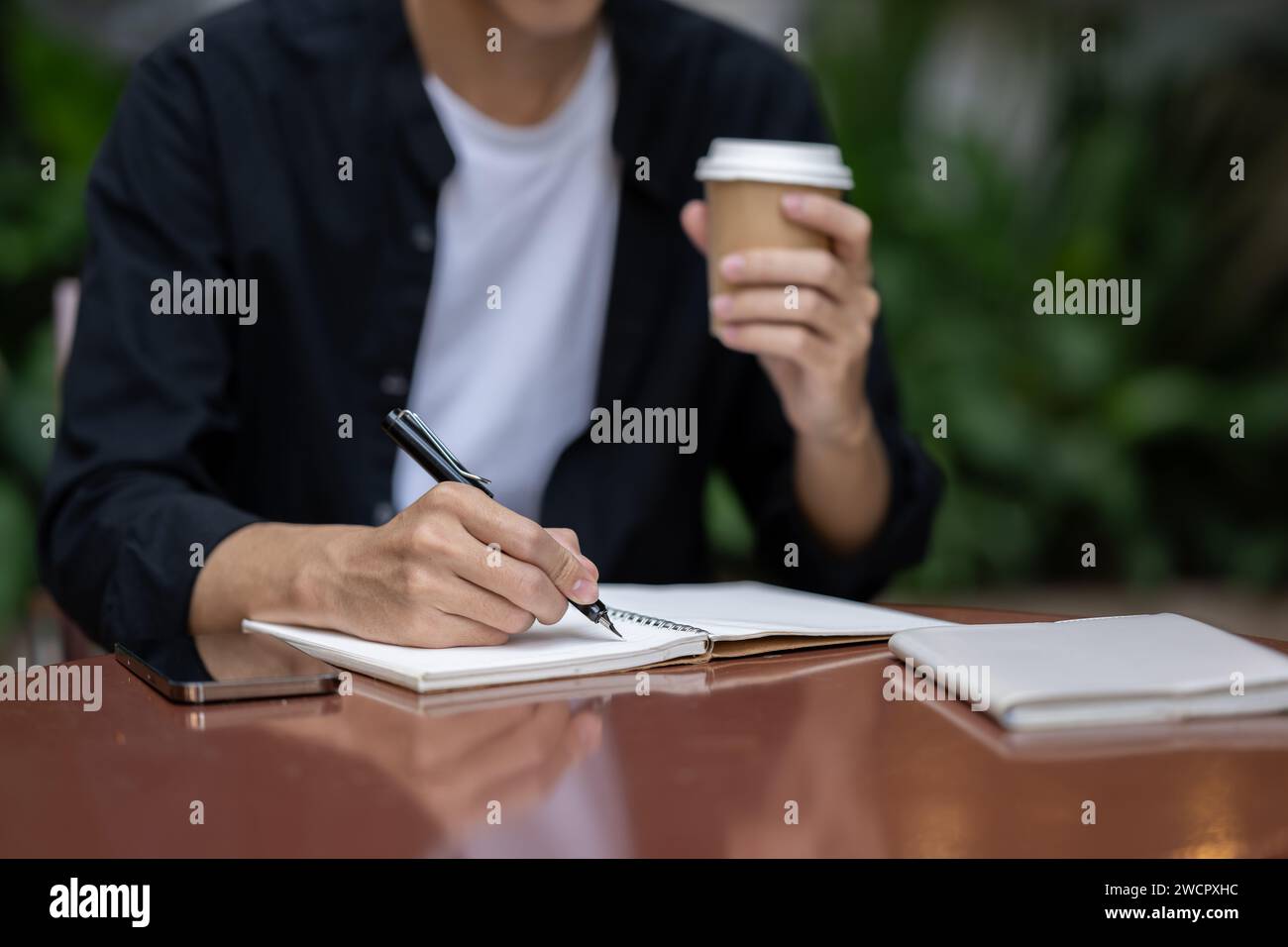 Close-up image of a young Asian man writing something in his book and sipping coffee at a table in a coffee shop. working remotely, digital nomad, fre Stock Photo