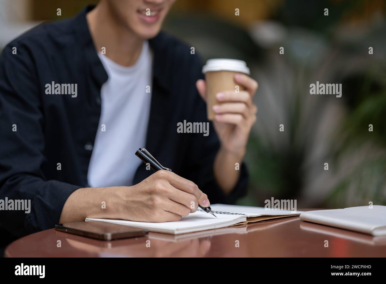 Close-up image of a young Asian man writing something in his book and sipping coffee at a table in a coffee shop. working remotely, digital nomad, fre Stock Photo