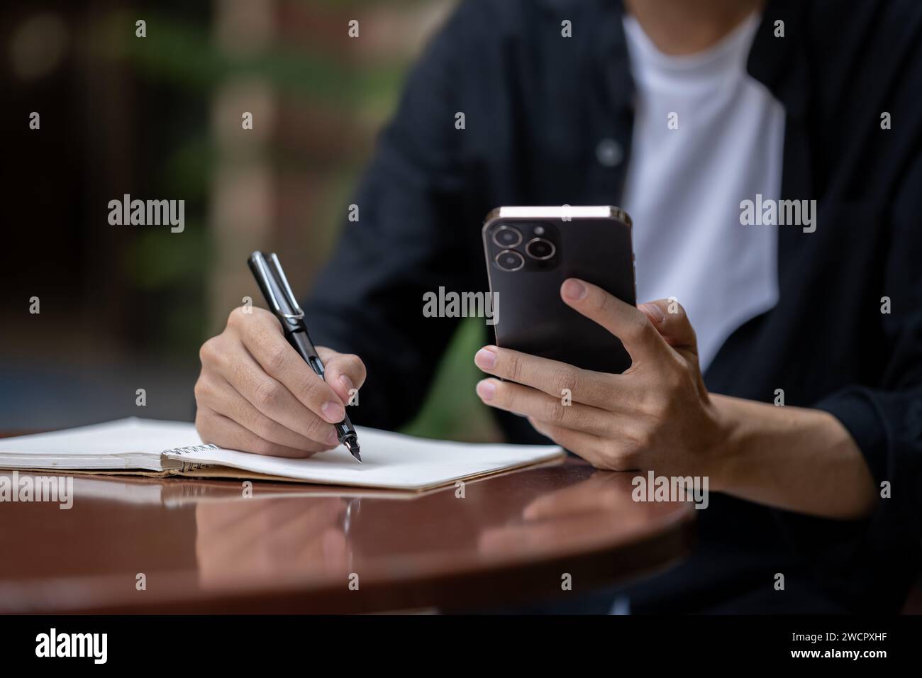 Close-up image of a man using his smartphone and taking notes in his book at a table in a coffee shop. Lifestyle concept Stock Photo