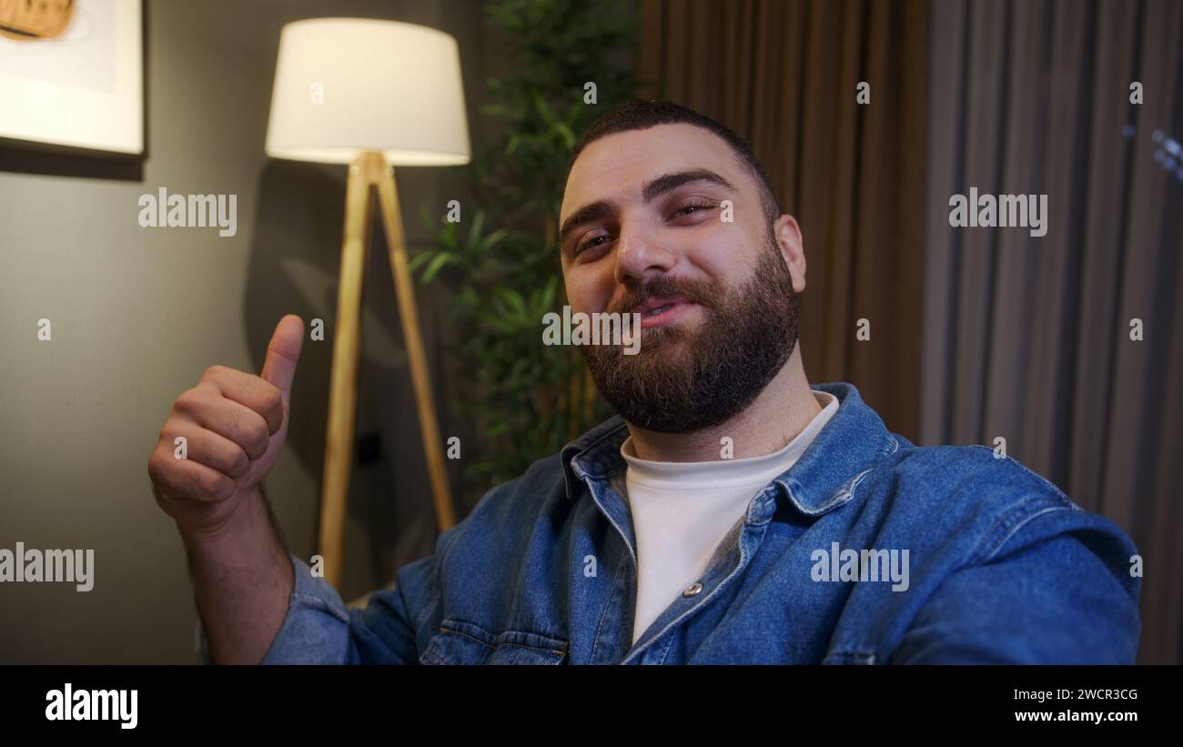 Happy young influencer adult man holding smartphone in hand making live video or vlog while sitting on sofa at home at night. Having fun, enjoy, leisu Stock Photo