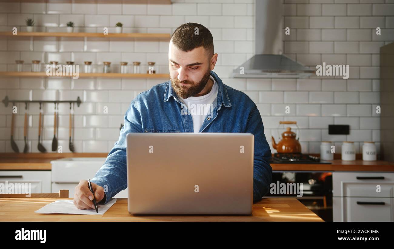 Bearded young adult man wearing in-ear headphones typing on laptop doing research sitting against the kitchen working on laptop Stock Photo