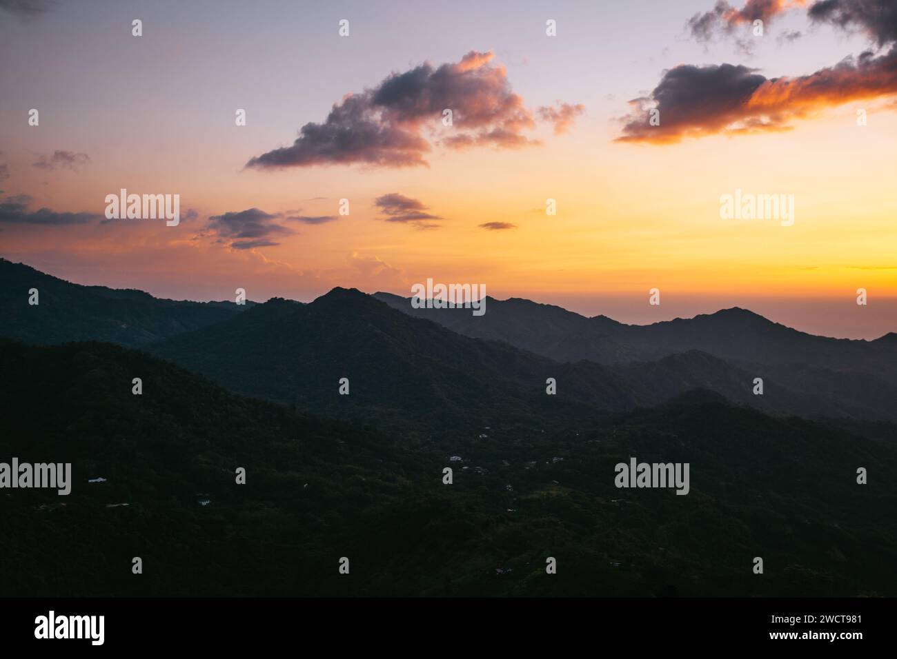 A tranquil sunset over layered mountain peaks with a warm, orange-toned sky and scattered clouds Stock Photo