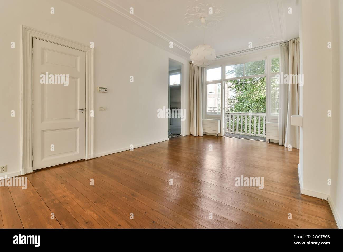 An empty room featuring polished hardwood floors, white walls, and large windows with light drapes. The space is adorned with a simple ceiling light a Stock Photo