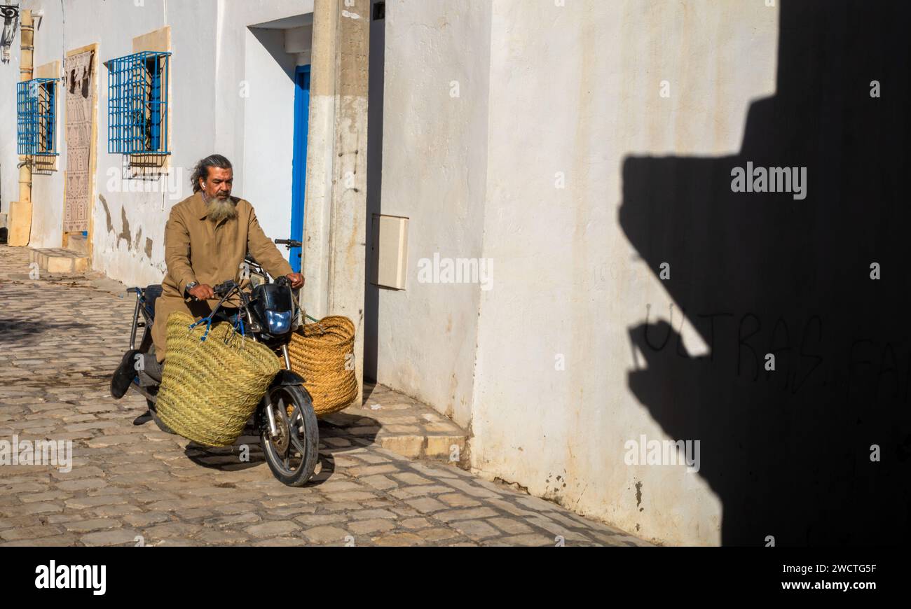 A bearded Tunisian man wearing earpods  carries large hand woven bags on his motorbike through a cobbled lane in the ancient medina of Sousse in Tunis Stock Photo