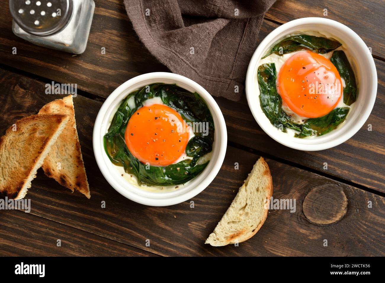 Baked eggs with vibrant orange yolks with cream, сheese and spinach in portion bowl served with grilled bread slices for dipping. Eggs en cocotte in r Stock Photo