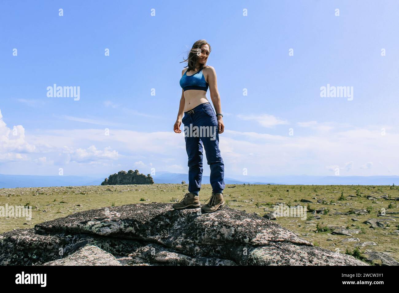 Woman tourist with good figure in blue pants and sports bra stands on top of cliff. Green meadow with stones, rock in distance background. Natural lan Stock Photo