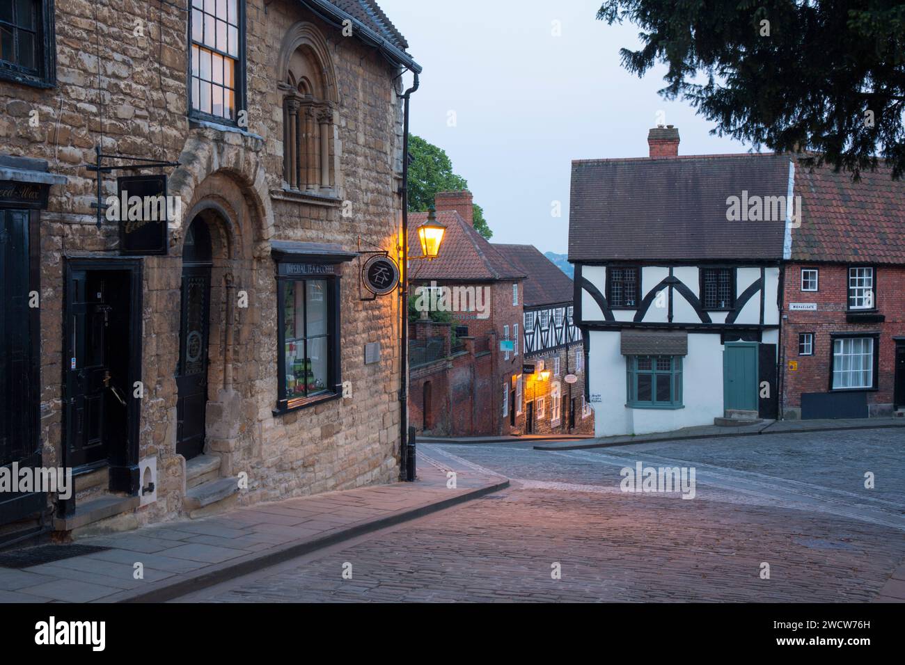 Lincoln, Lincolnshire, England. View down cobbled Steep Hill at the junction with Michaelgate, dusk, street lamps illuminating historic buildings. Stock Photo